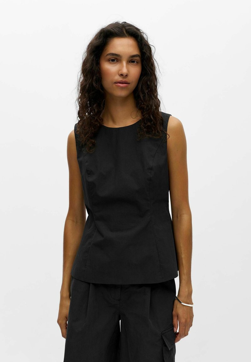 Femme aux cheveux bouclés foncés portant un haut noir sans manches et un pantalon noir assorti sur un fond blanc uni.