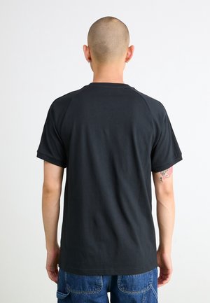 Mand med barberet hoved iført sort kortærmet t-shirt og blå jeans, stående med ryggen mod kameraet foran en ensfarvet hvid baggrund.