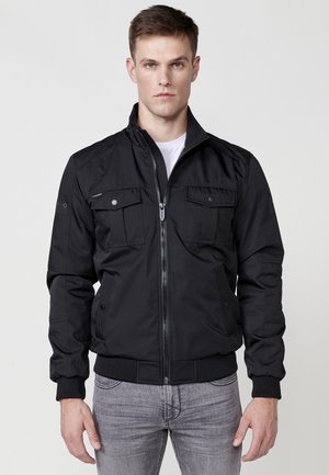 Chaqueta bomber negra con cremallera frontal, dos bolsillos en el pecho y puños acanalados. El material parece suave y con un ligero brillo.