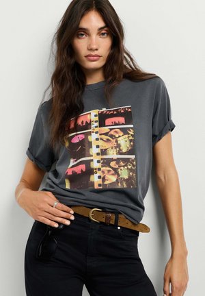 Vrouw met lang donker haar die een donker grafisch T-shirt met opgestroopte mouwen draagt, zwarte broek en een bruine riem met een zonnebril die uit de zak hangt.