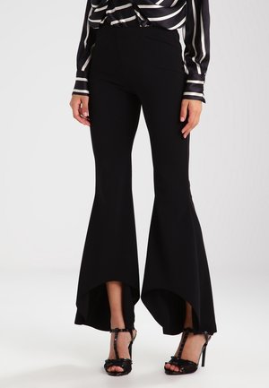 Frau trägt schwarze High-Low-Schlaghosen kombiniert mit schwarzen Sandalen mit hohem Absatz, steht vor einem schlichten Hintergrund.