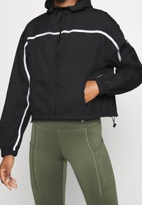 Veste courte noire avec capuche, dotée de bandes réfléchissantes blanches, fermeture éclair et poignets élastiques, associée à des leggings vert olive.