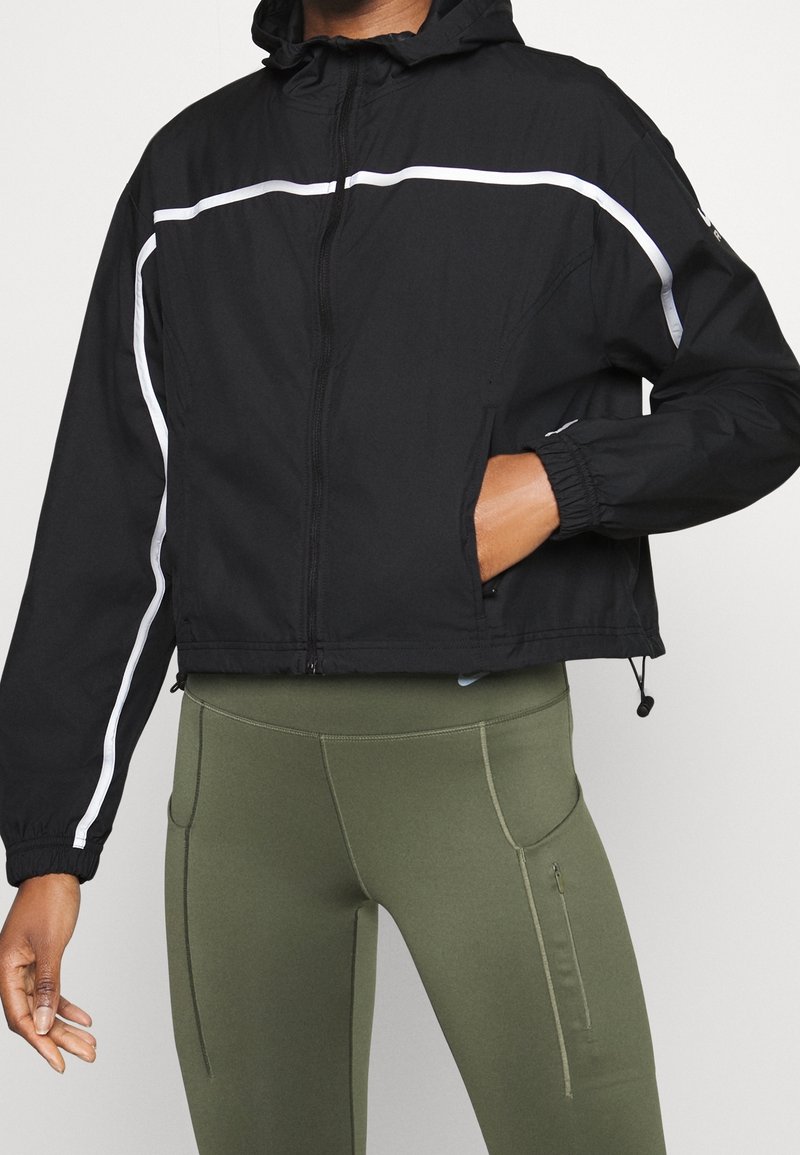 Veste courte noire avec capuche, dotée de bandes réfléchissantes blanches, fermeture éclair et poignets élastiques, associée à des leggings vert olive.