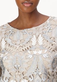 Beige brodert bluse med intrikate sølvblomsterdesign, transparent tekstur og rund halsutskjæring.