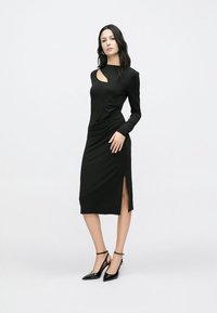 Svart midikjole med lange ermer, sidesplitt og en utskjæring ved halsen. Jevn tekstur; kombinert med svarte høye hæler.