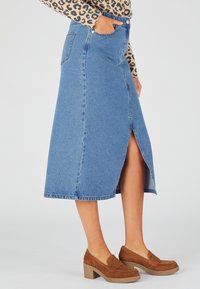 Falda midi de mezclilla en azul medio con una abertura frontal, diseño de cinco bolsillos y cintura ajustada. Combinada con zapatos de tacón cuadrado de ante marrón.