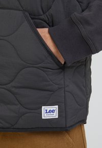 Steppad grå jacka med ribbad mudd, utrustad med en ficka och en etikett som säger "Lee Workwear". Tyget har en slät yta och en subtilt mönster.