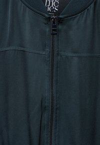 Mørkegrøn bomberjakke med lynlåslukning foran, ribbet krave og glat tekstur. Har kontrastfarvede syninger langs sømmene.