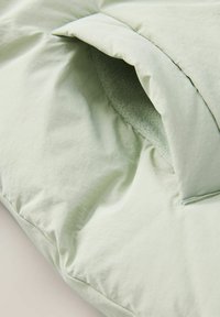 Vaaleanvihreä puhvitakki, jossa on pehmeä, teksturoitu vuori, hieman pöhöttyneet ulkopinnat ja vetoketjutasku sauman varrella.