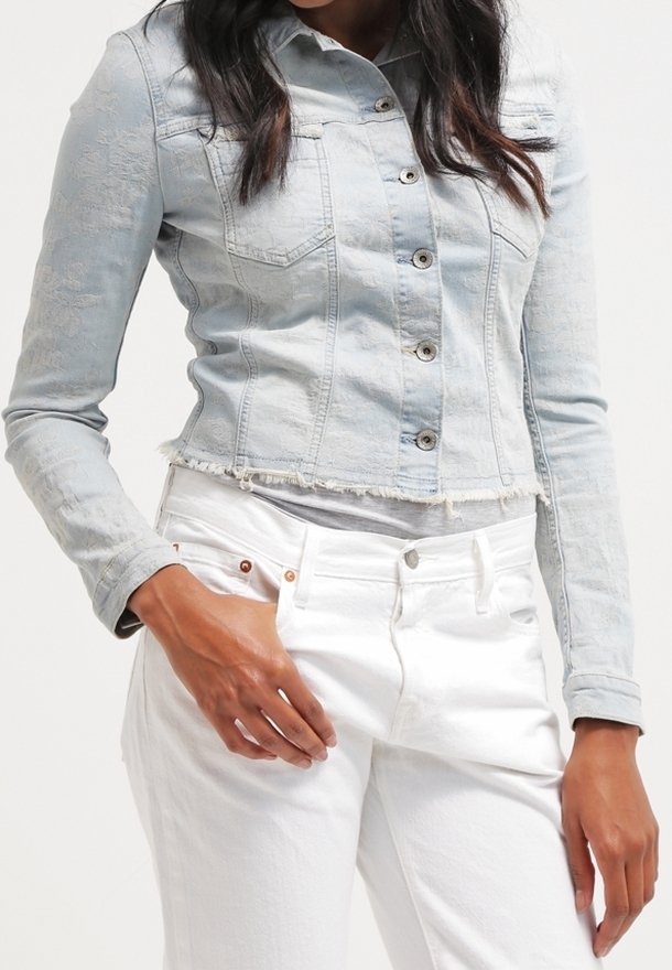 Donna che indossa una giacca di jeans cropped di colore azzurro chiaro con chiusura a bottoni e pantaloni bianchi a vita alta su uno sfondo neutro.