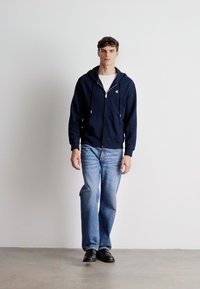 Hanorac navy cu fermoar și șnururi, purtat peste un tricou alb. Asortat cu blugi din denim de un albastru deschis și pantofi negri. Design simplu și croială confortabilă.