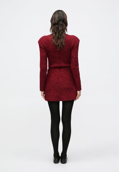 Červené pletené šaty s textúrou, s vypchávkami na ramenách, priliehavým pásom a krátkou dĺžkou. Nosia sa s čiernymi pančuchami a lodičkami na podpätku, pohľad zozadu.