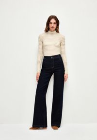 Hvid struktureret langærmet top med vandrette striber, parret med højtaljede mørkeblå flared jeans og beige slip-on sko.