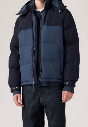Homme portant une doudoune d'hiver bicolore bleu marine et bleu foncé avec une fermeture éclair devant et des poignets à boutons-pression.