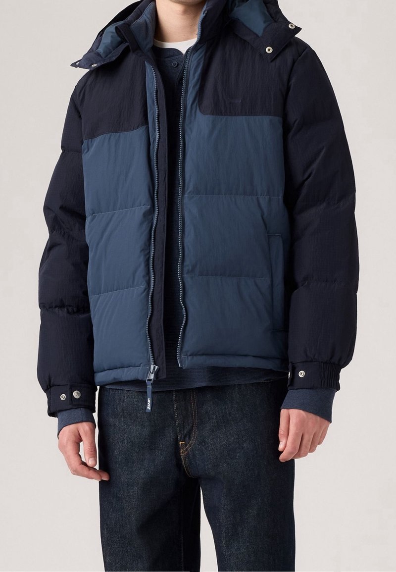 Homme portant une doudoune d'hiver bicolore bleu marine et bleu foncé avec une fermeture éclair devant et des poignets à boutons-pression.