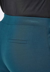 Teal high-waisted broeken met een gladde textuur, met een klein achterzakje en een platte tailleband, gedeeltelijk zichtbaar naast een zwarte top.