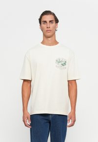 Ung mann med kort bølgete hår har på seg en kremfarget T-skjorte med fuglillustrasjon og teksten "The Gant Birding Club" samt blå jeans, stående foran en hvit bakgrunn.