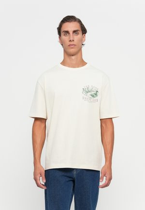 Ung mann med kort bølgete hår har på seg en kremfarget T-skjorte med fuglillustrasjon og teksten "The Gant Birding Club" samt blå jeans, stående foran en hvit bakgrunn.