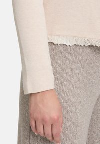 Pull léger rose avec une finition texturée et un ourlet effiloché, associé à un pantalon à motif chevron beige. Main reposant le long de la couture.