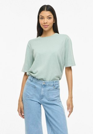 Ung kvinde med langt mørkt hår iført en lysegrøn kortærmet skjorte indtuckt i højtsiddende lyseblå jeans, stående med afslappet kropsholdning.