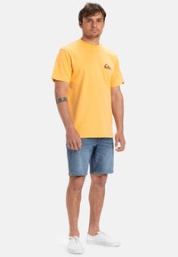Camiseta de algodón amarilla con mangas cortas y un pequeño logo en la parte superior izquierda. Combinada con pantalones cortos de mezclilla y zapatillas blancas.