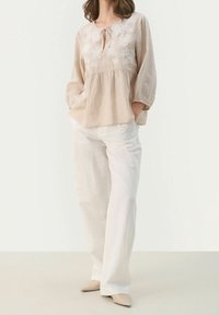 Beige pellavapusero, jossa kukkakirjailu, väljä istuvuus, pyöreä pääntie nauhoilla, 3/4-hihat; yhdistetty valkoisiin pellavahousuihin. Neutraalinsävyiset kengät.