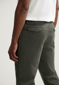 Donkergroene corduroy broek met een slim fit, met achterzakken en een knoopdetail, zijwaarts en vanaf de taille gezien tegen een neutrale achtergrond.