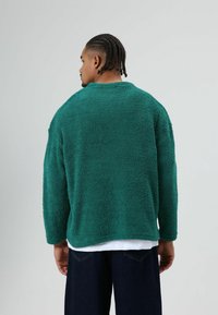 Turkooise, oversized trui met een textuur, een hoge hals en lange mouwen. Draagt over een wit shirt, gecombineerd met donkerblauwe denim jeans.