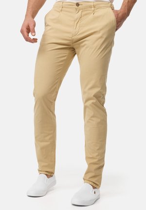 Beige chino pants made of cotton blend, slim fit design with front pockets, zip fly, and a single button closure. Paired with white slip-on shoes.