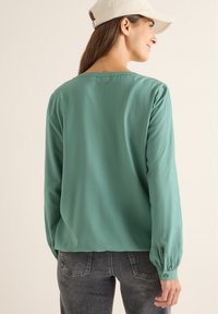 Langärmlige, türkise Bluse mit fließender Textur, rundem Ausschnitt und geknöpften Manschetten, kombiniert mit grauen tapered Jeans.