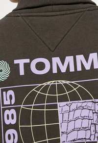 Felpa marrone con la scritta "TOMMY" in viola chiaro, grafica circolare e design del globo. Presenta un colletto a coste e un dettaglio di pattern testurizzato unico.