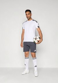 Vīrietis baltā futbola kreklā, tumšās šortos un baltās zeķēs tur futbolu bumbu pret vienkāršu gaišo fona.