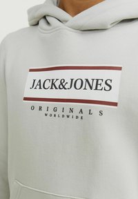 Lichtgrijze hoodie met een grote voorkantgrafiek met de tekst "JACK&JONES" en "ORIGINALS WORLDWIDE" in zwart en bordeauxrood. Zachte stoftextuur.