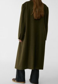 Olijfgroene wollen jas met een lange, losse pasvorm, wijde mouwen en zijzakken, gecombineerd met donkere spijkerbroeken en casual bruine schoenen.