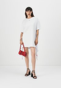Bílé oversized tričkové šaty s oblým lemem, v kombinaci s malou červenou kabelkou se květinovým vzorem a černými sandály na podpatku.