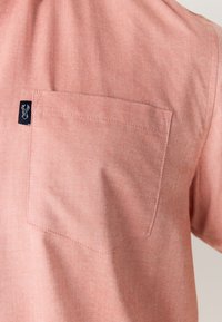 Kortærmet skjorte i lys koralfarvet stof, med brystlomme og et lille sort logomærke på lommens søm. Glat tekstur.