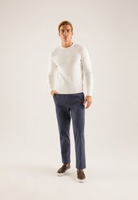 Pull en maille blanche, pantalon bleu marine, chaussures marron sans lacets. Le mannequin se tient devant un fond neutre, présentant une tenue décontractée.