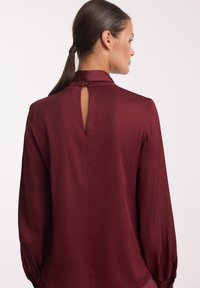 Bordowa bluzka z wysokim kołnierzem, długimi rękawami i detalem w kształcie klucza z tyłu. Gładka tekstura, dopasowana w talii, elegancki układ.