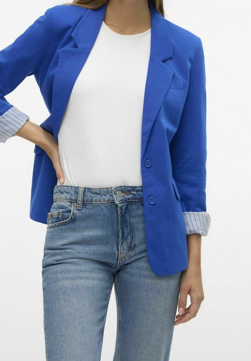 Niebieska marynarka o gładkiej fakturze, z wciętymi klapami i zapięciem na guziki. W połączeniu z białą koszulą i mankietem w jasnoniebieskie paski. Dopasowane dżinsy.