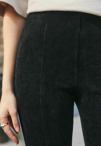Detailní záběr na osobu v černých vysokých ribkovaných kalhotách a béžovém topu, s rukou, která ukazuje zelený kamenný prsten.