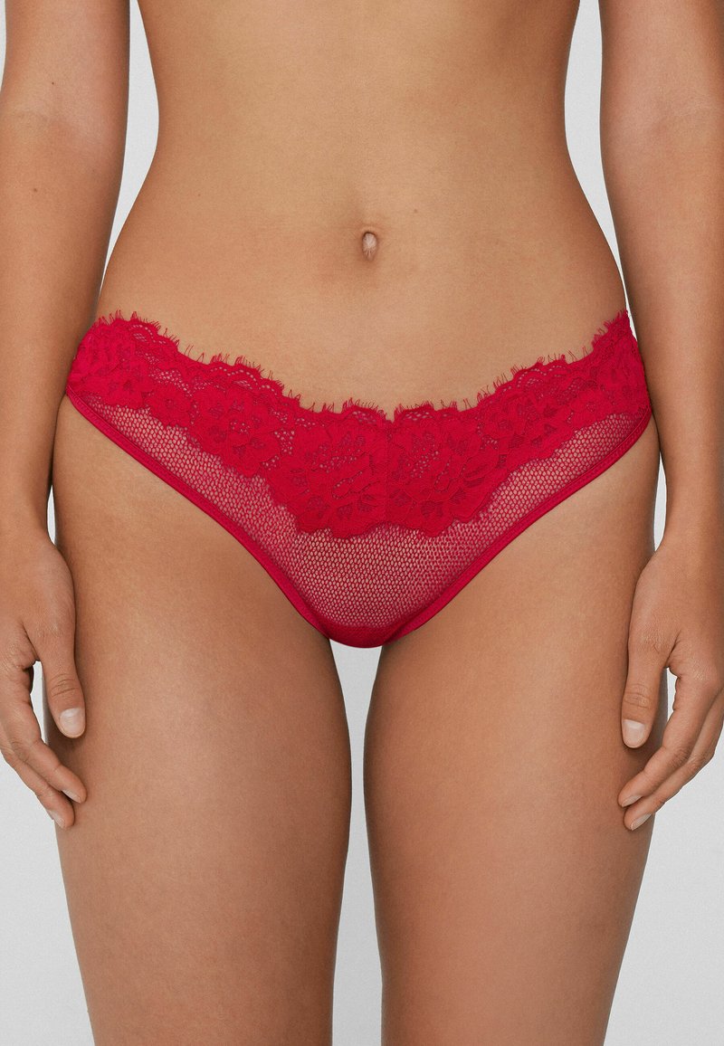 String en dentelle rouge avec motif floral à la taille, doté d'un corps en résille et d'une texture douce. Coutures plates et absence de ferrures visibles.