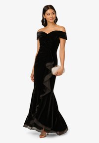 Zwarte fluwelen off-shoulder jurk met een nauwsluitend lijfje, gerimpelde zoom en zij draping. Model houdt een zilveren clutch vast; draagt sandaaltje met bandjes.