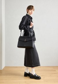 Czarna skórzana torebka o strukturalnym kształcie, z przednią klapą i srebrnymi elementami. Model nosi dżinsową kurtkę oraz ciemną spódnicę midi z trampkami.