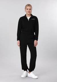 Jeune femme debout, portant un sweat à capuche noir zippé et un pantalon de jogging assorti avec des baskets blanches, sur un fond gris uni.