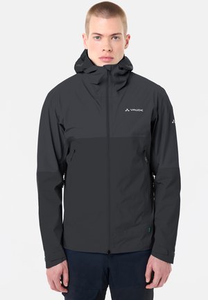 Junger Mann trägt eine schwarze Vaude Kapuzenjacke mit Reißverschluss, steht vor einem schlichten hellen Hintergrund und blickt in die Kamera.