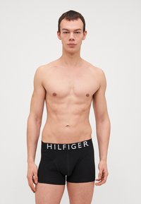 Fekete boxer alsónadrág elasztikus derékkal, amelyen fehér "HILFIGER" logó található. Sima textúra, testhezálló kialakítás, combközép hosszúság.