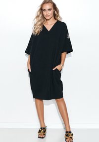 Czarna, oversize sukienka z dekoltem w serek, krótkimi, szerokimi rękawami, bocznymi kieszeniami i luźnym krojem. Posiada akcent logo na rękawie.