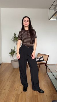 Jeune femme aux longs cheveux noirs portant un polo rayé marron et un pantalon large noir, debout dans une pièce moderne avec un sol en bois.