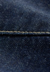 Denimstof i mørkeblå med en glat tekstur, der har synlige væve mønstre og en markant tan syning langs kanten.