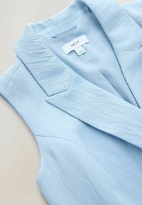 Blazer bleu clair texturé avec col à revers cranté et étiquette de la marque "NEXT" visible à l'intérieur du col.